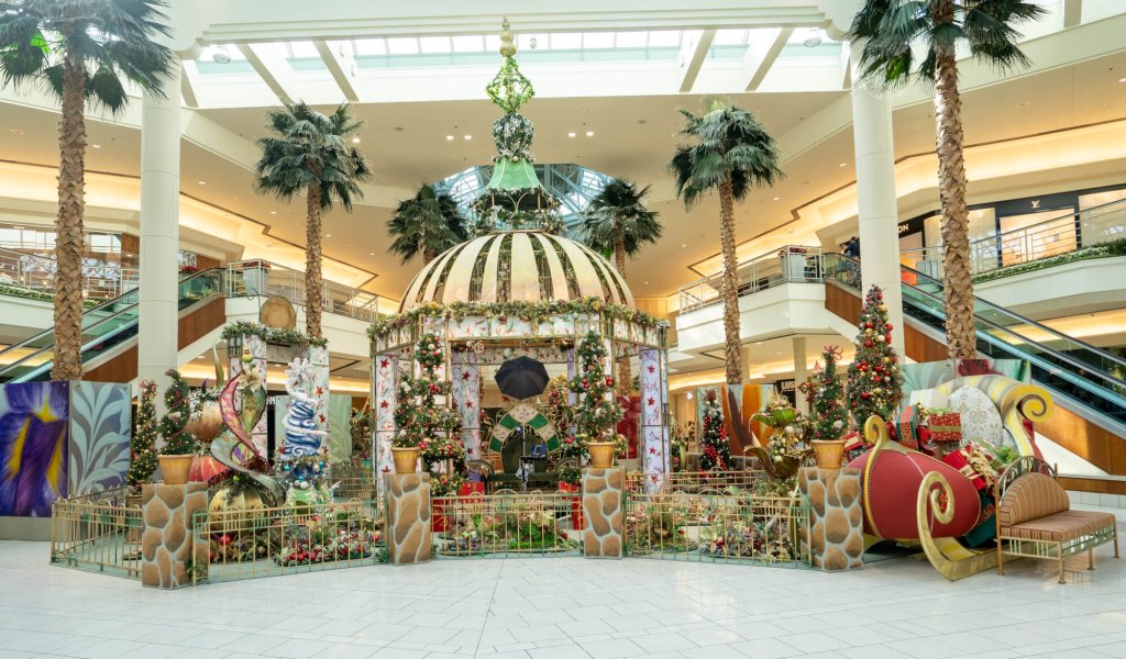A festive holiday display featuring a golden sleigh, Christmas trees, and a large dome adorned with lights and decorations in a shopping mall atrium