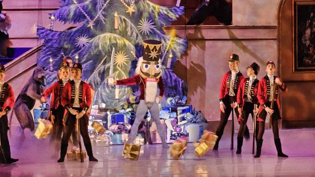 A festive ballet performance featuring a Nutcracker soldier and a mouse king battling amidst a backdrop of a decorated Christmas tree and gift boxes