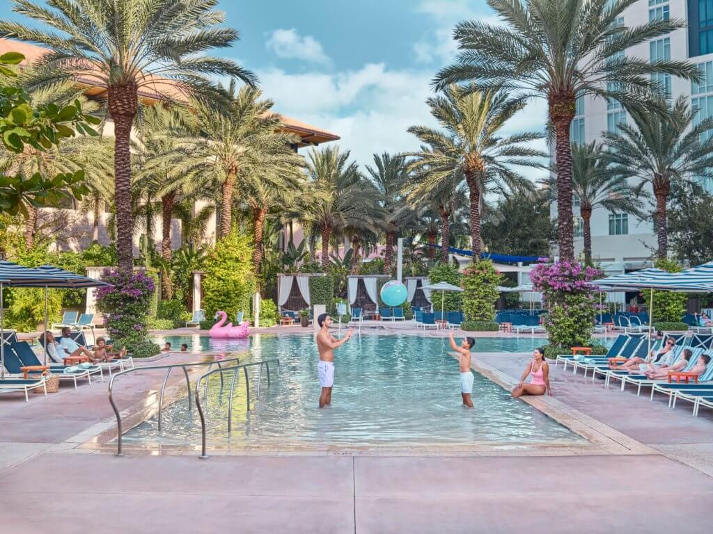 A vibrant resort pool scene with palm trees, colorful flowers, and guests enjoying the water and surrounding lounge chairs