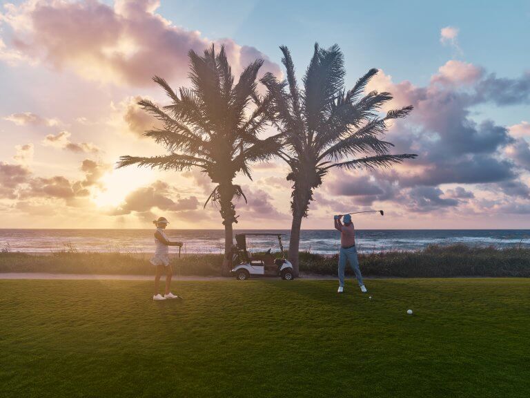 Dos golfistas disfrutan de una tranquila partida al atardecer en un campo de golf tropical, con palmeras y el océano de fondo