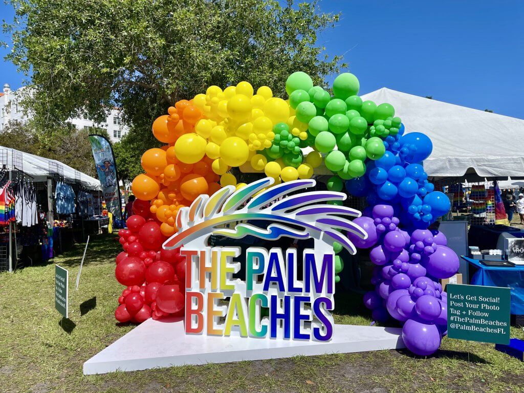 A vibrant rainbow balloon arch with the words 'The Palm Beaches' in colorful letters, set against a backdrop of white tents and green trees