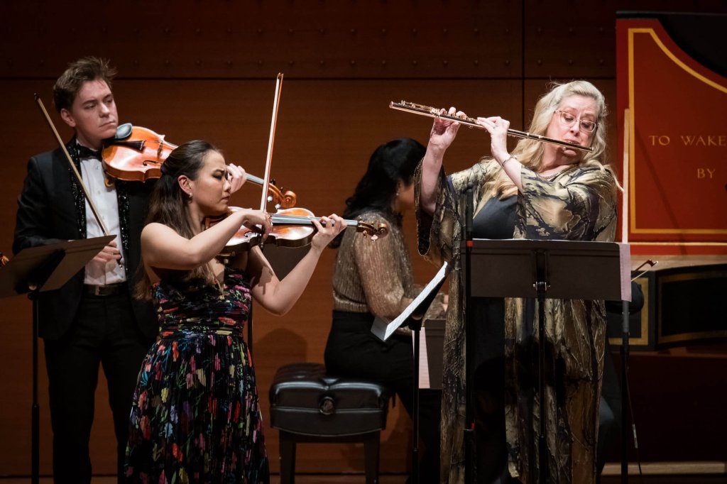 A string quartet performs on stage, with two violinists, a violist, and a cellist, all dressed in formal attire