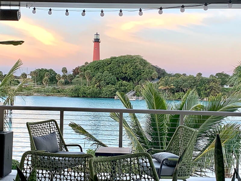 Eine ruhige Terrasse am Wasser mit Korbstühlen und einem Tisch mit Blick auf einen ruhigen See und einen malerischen Leuchtturm, der sich in der Ferne inmitten von üppigem Grün erhebt