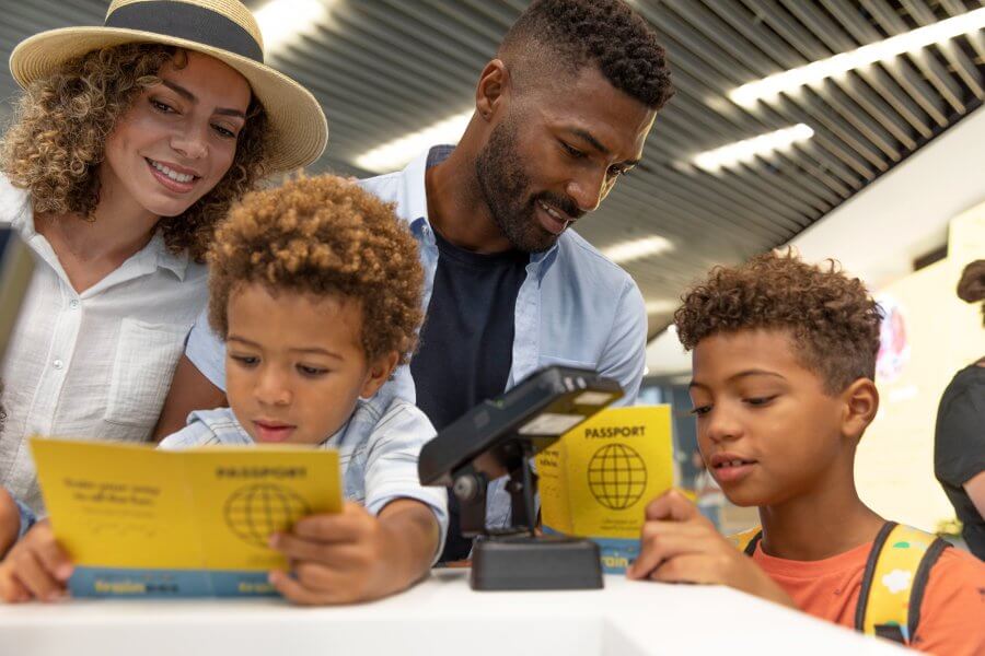 A family of four, including two young boys, is gathered around a passport control kiosk at an airport, with the children excitedly holding their yellow passports