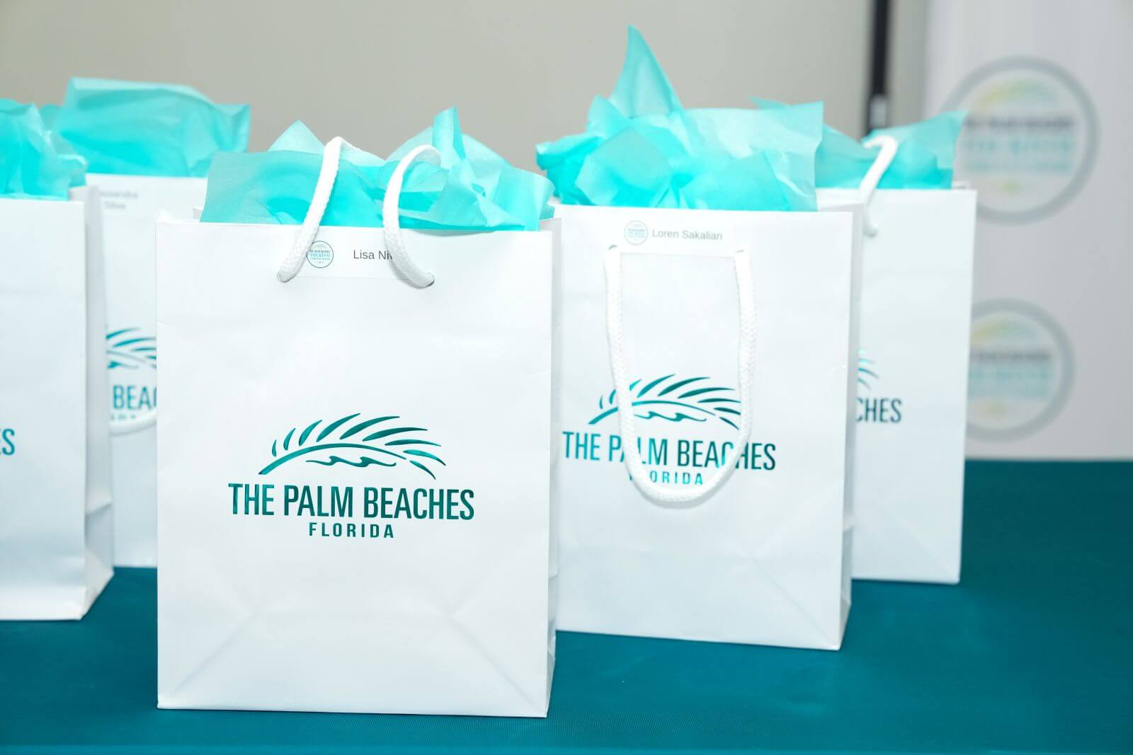 Four white gift bags with turquoise tissue paper peeking out, each featuring the logo and text 'The Palm Beaches, Florida'