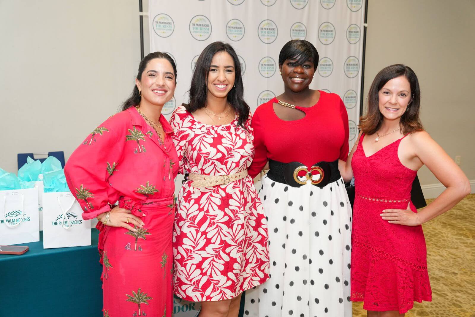 Four stylish women pose together in vibrant red dresses against a backdrop of white banners with circular logos