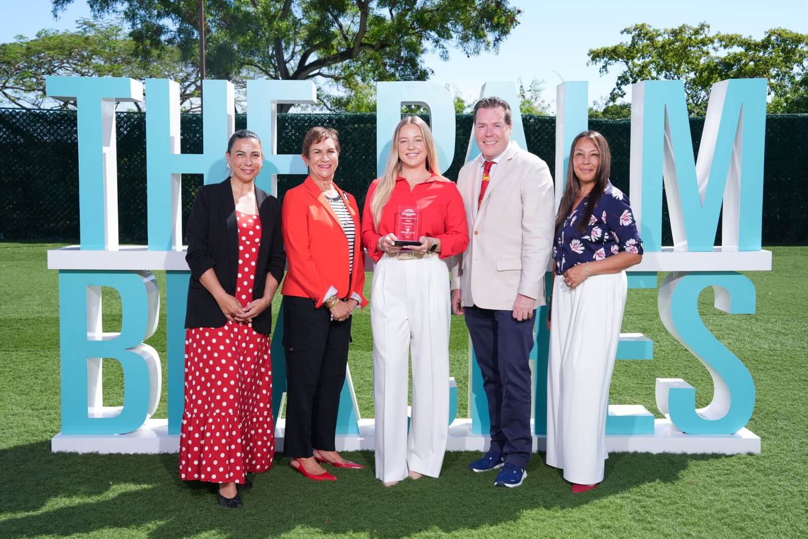 Five individuals stand together in front of a large sign reading 'The Palm Beach', smiling and dressed in formal attire