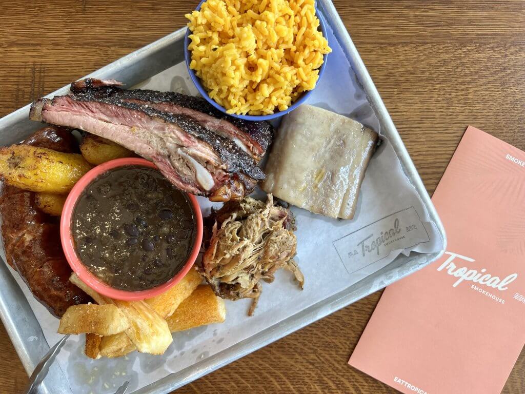 Una bandeja de metal contiene una variedad de carnes a la parrilla, como costillas, cerdo desmenuzado y salchichas, acompañadas de guarniciones de arroz amarillo, frijoles negros y plátanos