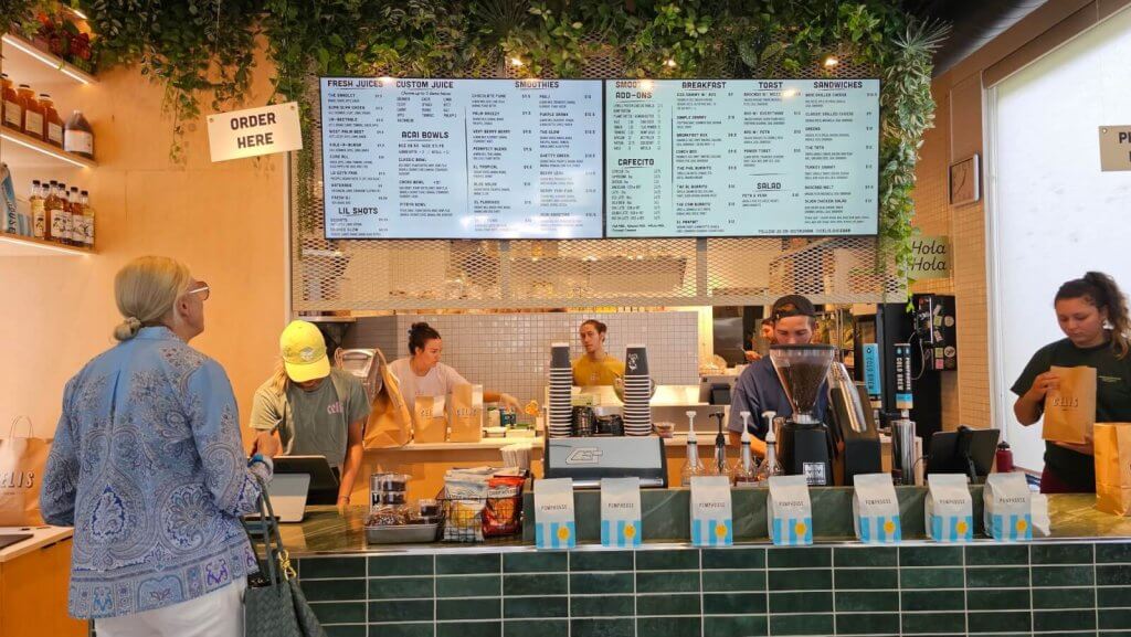 Una cafetería llena de gente con un cartel verde y llamativo colgado sobre el mostrador, donde los clientes hacen sus pedidos y el personal prepara bebidas y comida