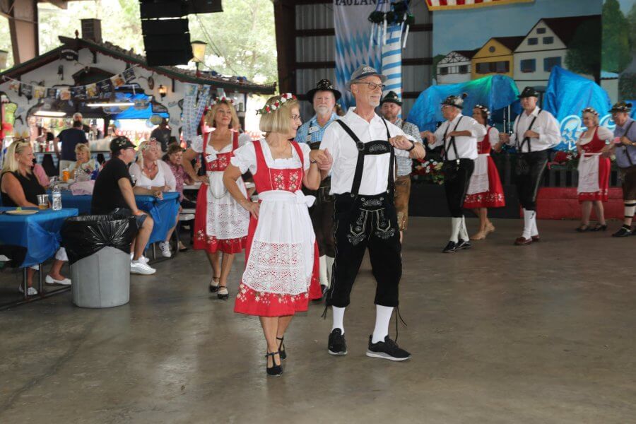 Spectacle de danse folklorique animé par des couples portant des costumes traditionnels bavarois, notamment des dirndls et des lederhosen, lors d'un festival en plein air avec des tentes et des décorations colorées.