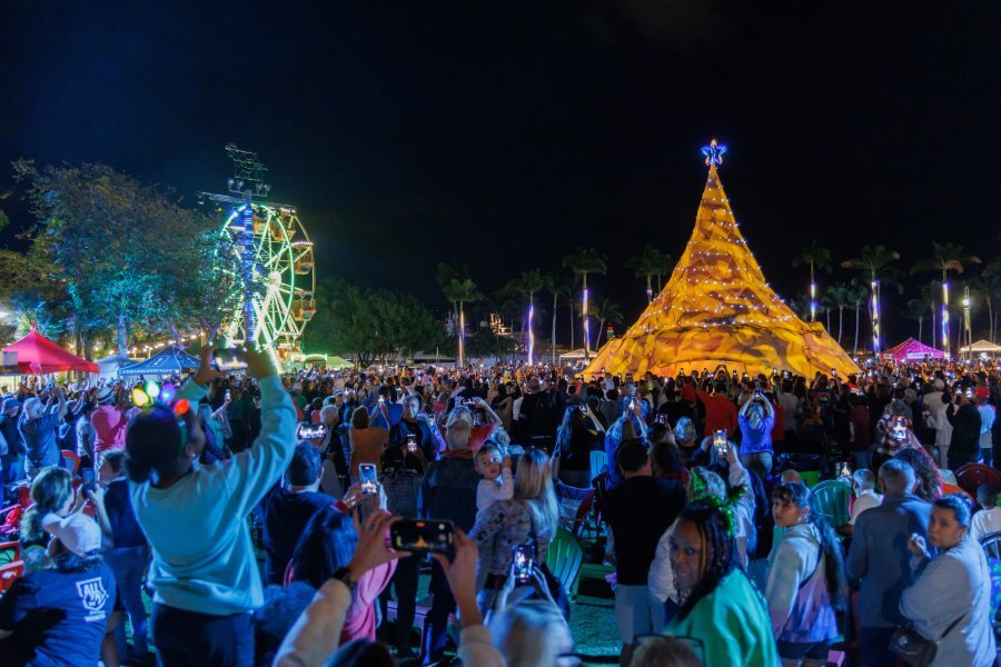 A festive night scene at a holiday fair with a large illuminated Christmas tree, a Ferris wheel, and a crowd of people enjoying the event