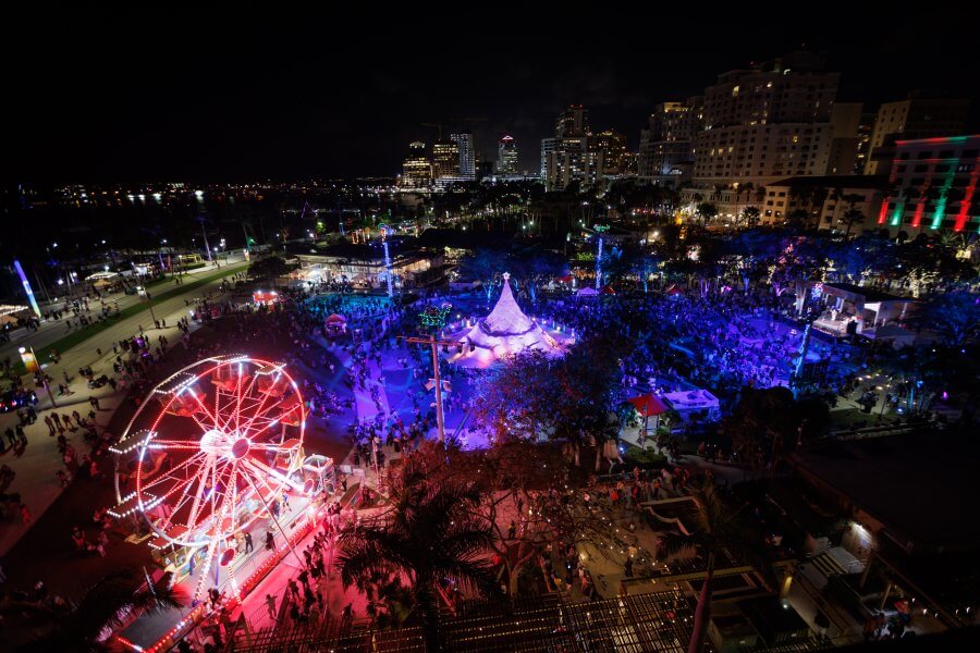 A vibrant night scene at a festive amusement park with a large crowd enjoying rides, games, and a spectacular Christmas tree illuminated by colorful lights