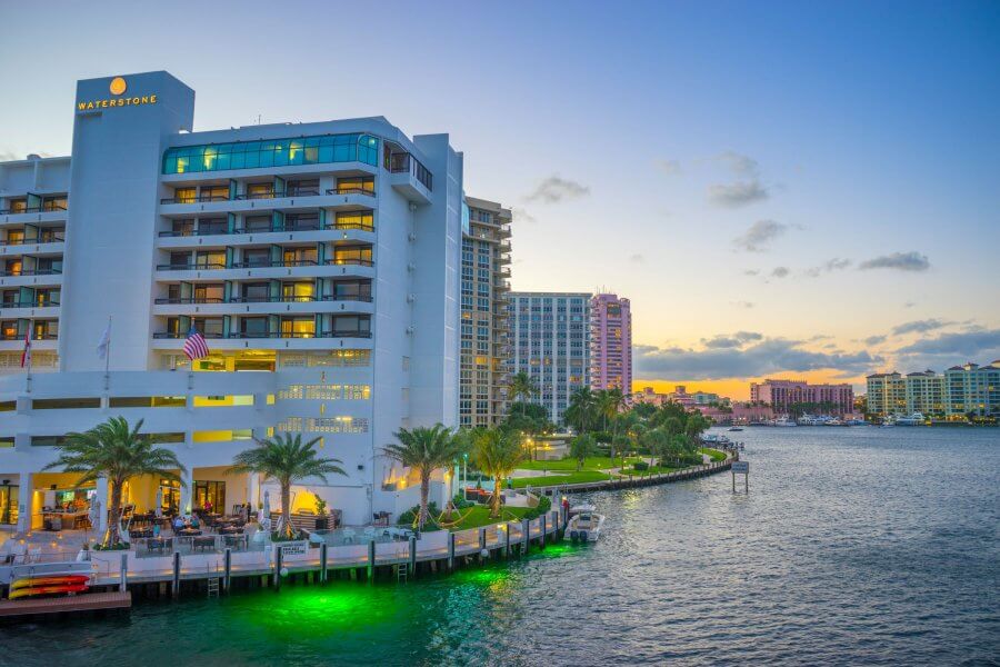 A serene waterfront scene at dusk featuring the Waterstone Hotel & Marina, surrounded by palm trees and illuminated by soft lights reflecting on the water