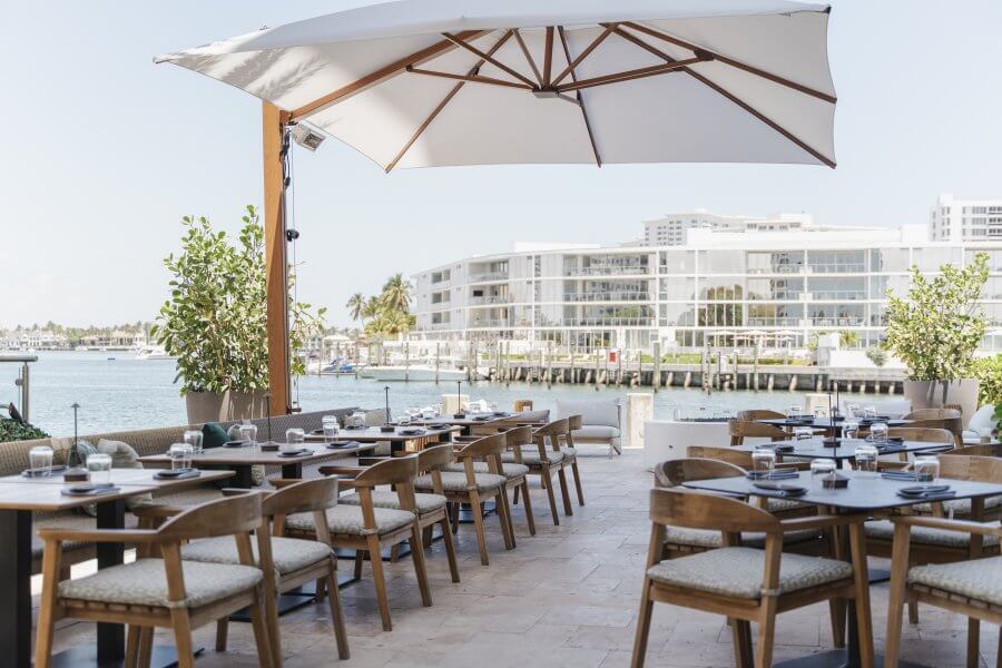 Outdoor waterfront restaurant with wooden tables and chairs under large umbrellas, overlooking a marina with boats and modern buildings in the background