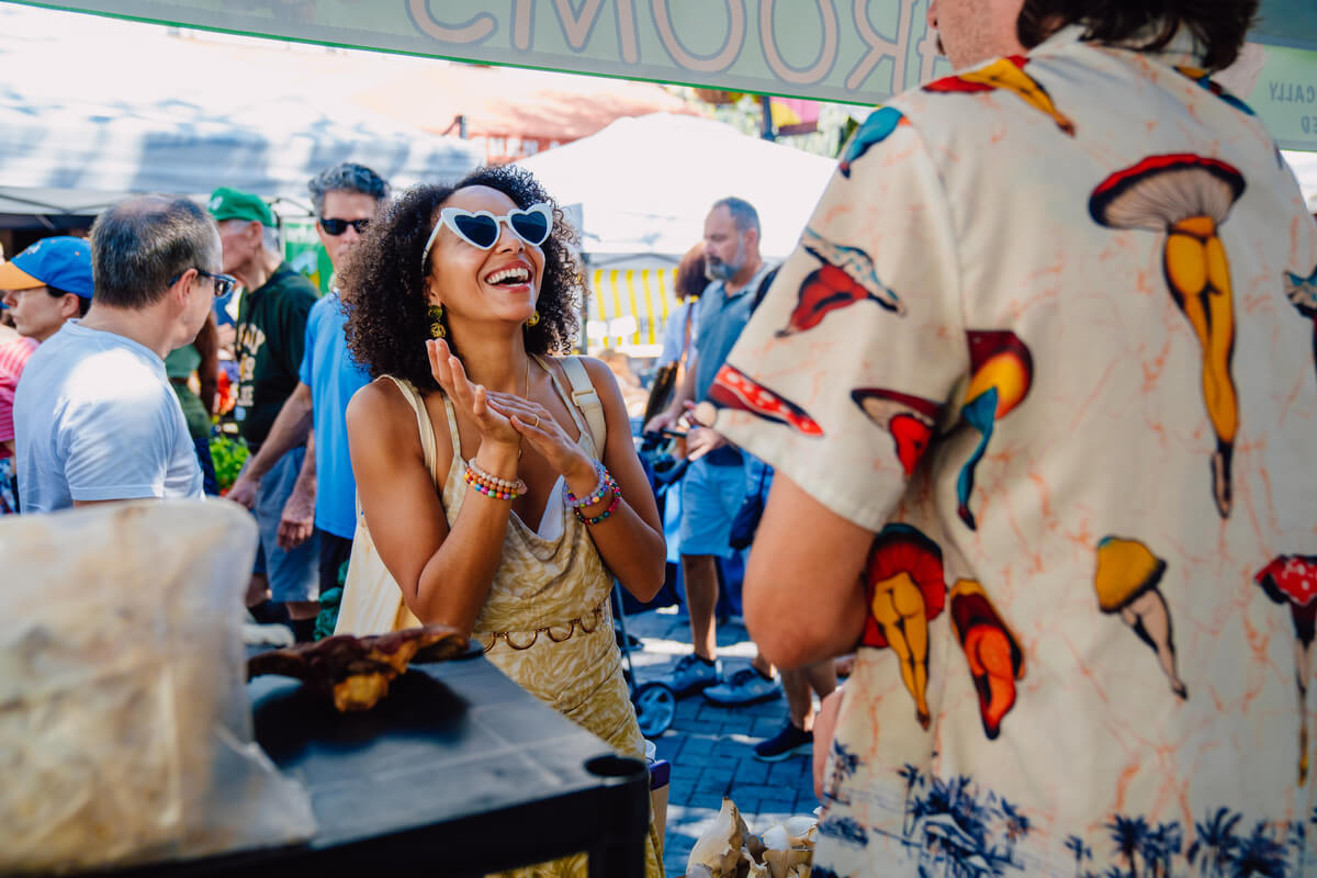 A woman wearing heart-shaped sunglasses laughs while talking to a person in a patterned shirt