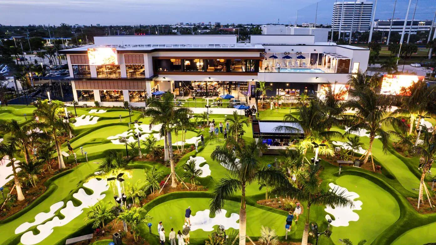 An aerial view of a vibrant, tropical mini-golf course with glowing green turf, surrounded by palm trees and a modern building complex with a rooftop pool and restaurant