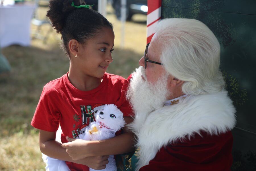 A young girl in a red Santa-themed shirt smiles as she holds a white stuffed animal next to Santa Claus, who is dressed in his traditional red and white outfit
