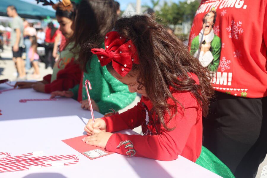 A young girl wearing a red dress and a large red bow in her hair is carefully writing a letter to Santa Claus at a holiday event