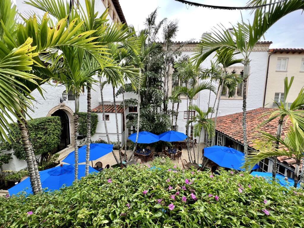 Un patio tropical con sombrillas azules y una exuberante vegetación, rodeado de edificios blancos con techos de tejas rojas