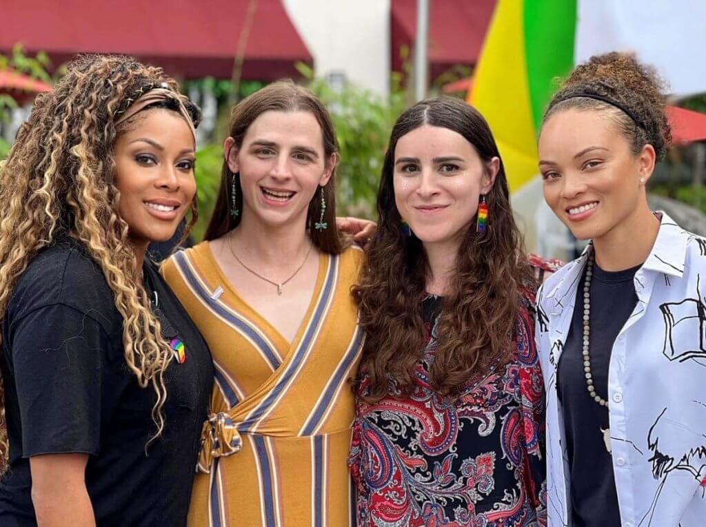 Four women with diverse hairstyles and outfits stand together outdoors, smiling and posing for a photo