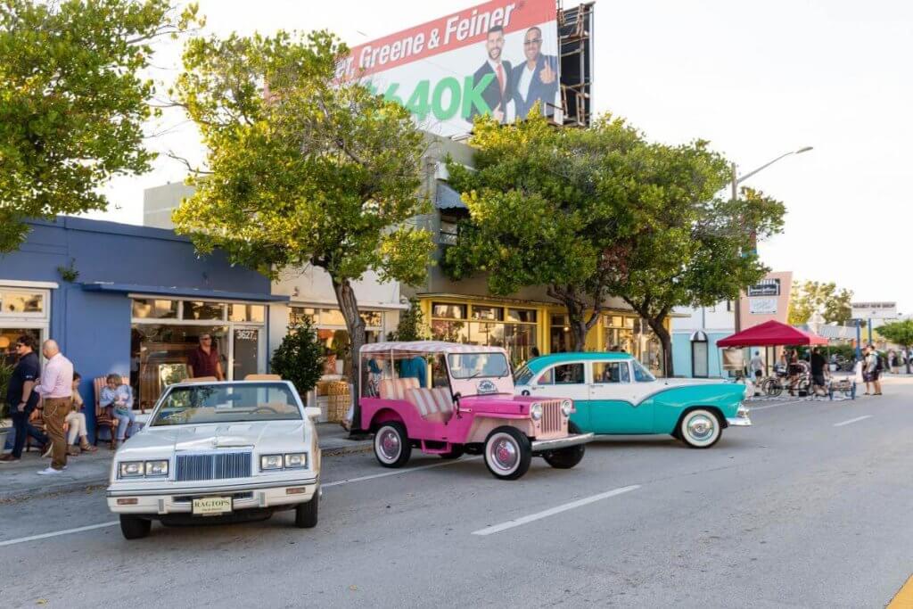 Vintage cars, including a white convertible, a pink jeep, and a turquoise classic, cruise down a palm-lined street in a vibrant neighborhood