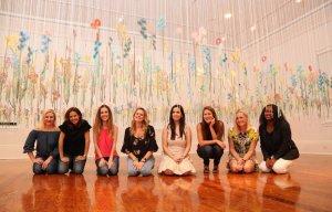 Un groupe de huit femmes posant ensemble dans une installation artistique colorée avec des décorations florales suspendues.