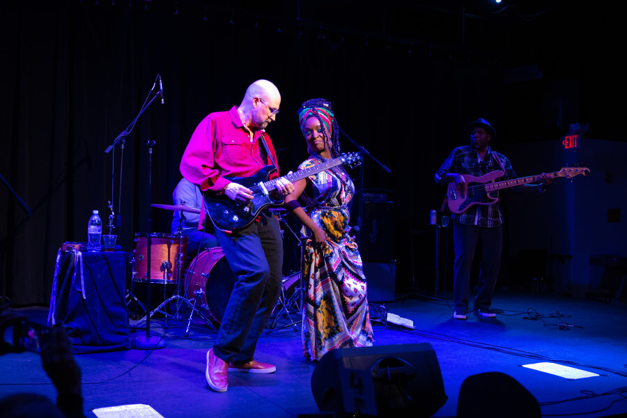 A vibrant musical performance featuring a diverse group of musicians on stage, with a guitarist and a drummer in the foreground and a bassist in the background