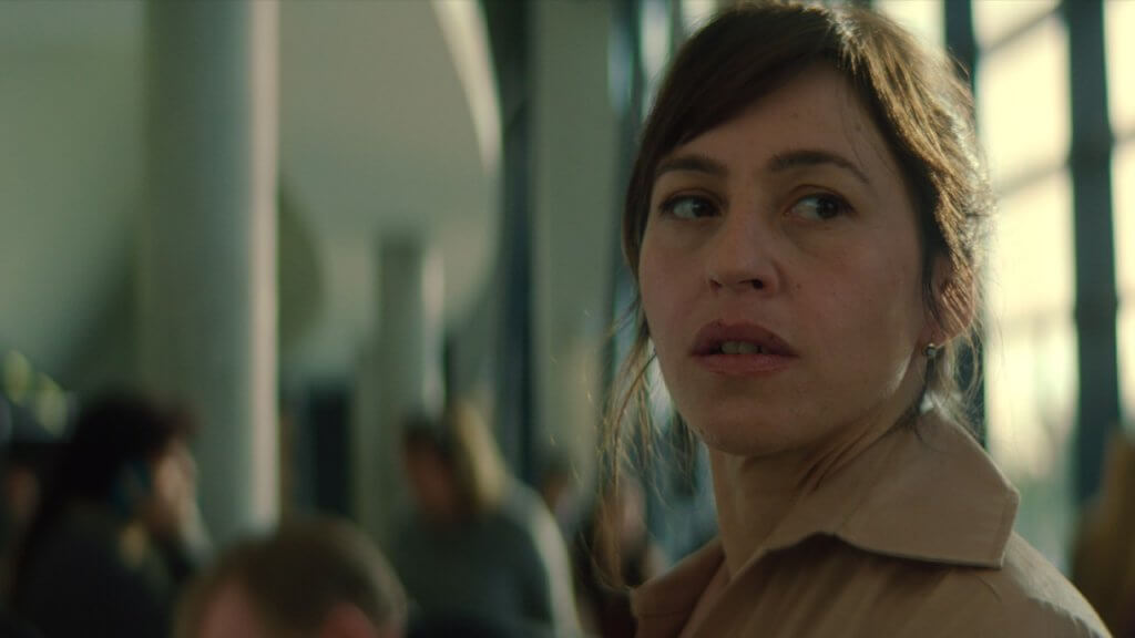 A woman with a concerned expression stands in a modern building with large windows and pillars