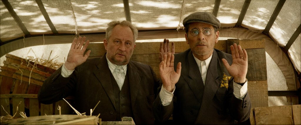 Two men in suits and hats raise their hands in a gesture of greeting or surrender inside a rustic wooden structure