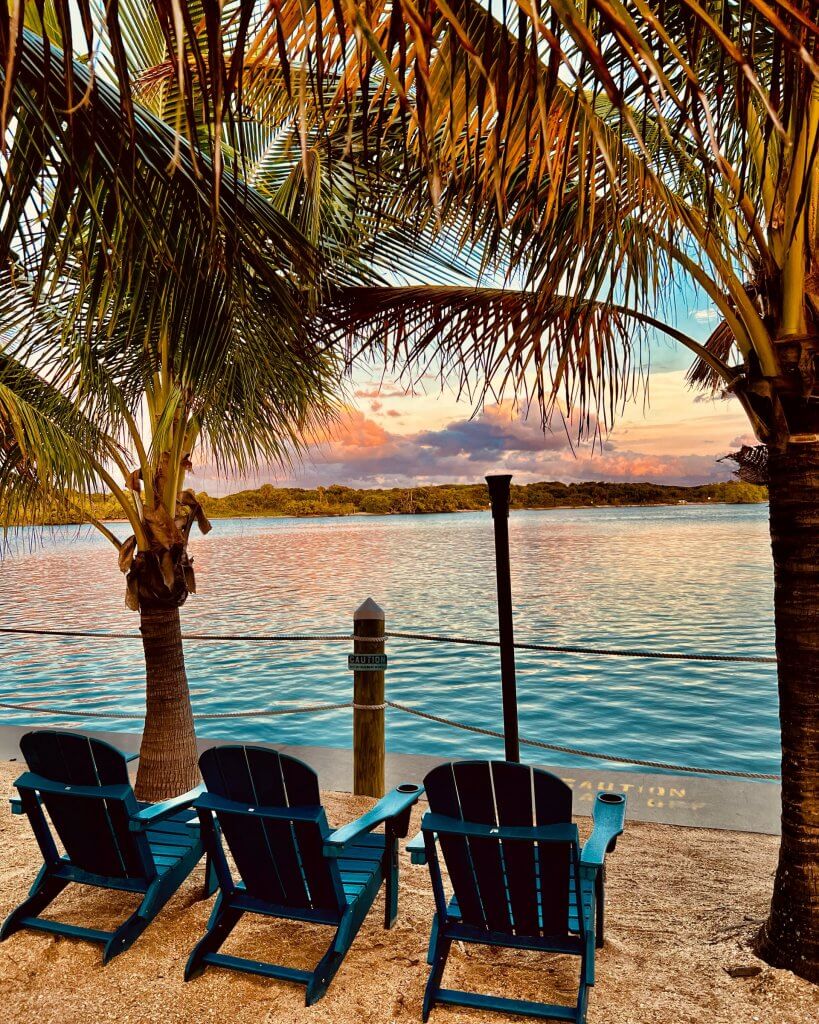 Tres sillas Adirondack azules dan a un lago tranquilo al atardecer, enmarcadas por palmeras y una barandilla de madera