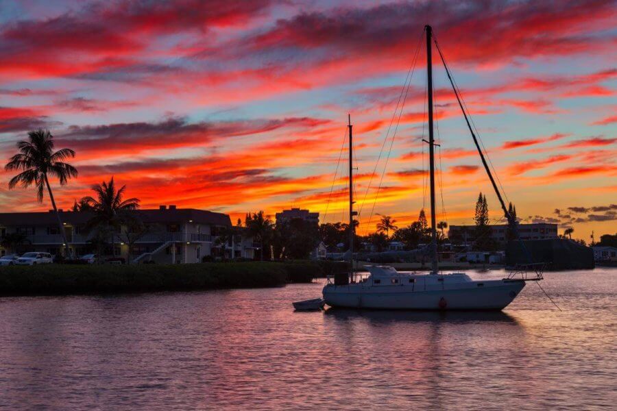 A serene sunset over a calm river with a sailboat gently floating, framed by palm trees and colorful clouds