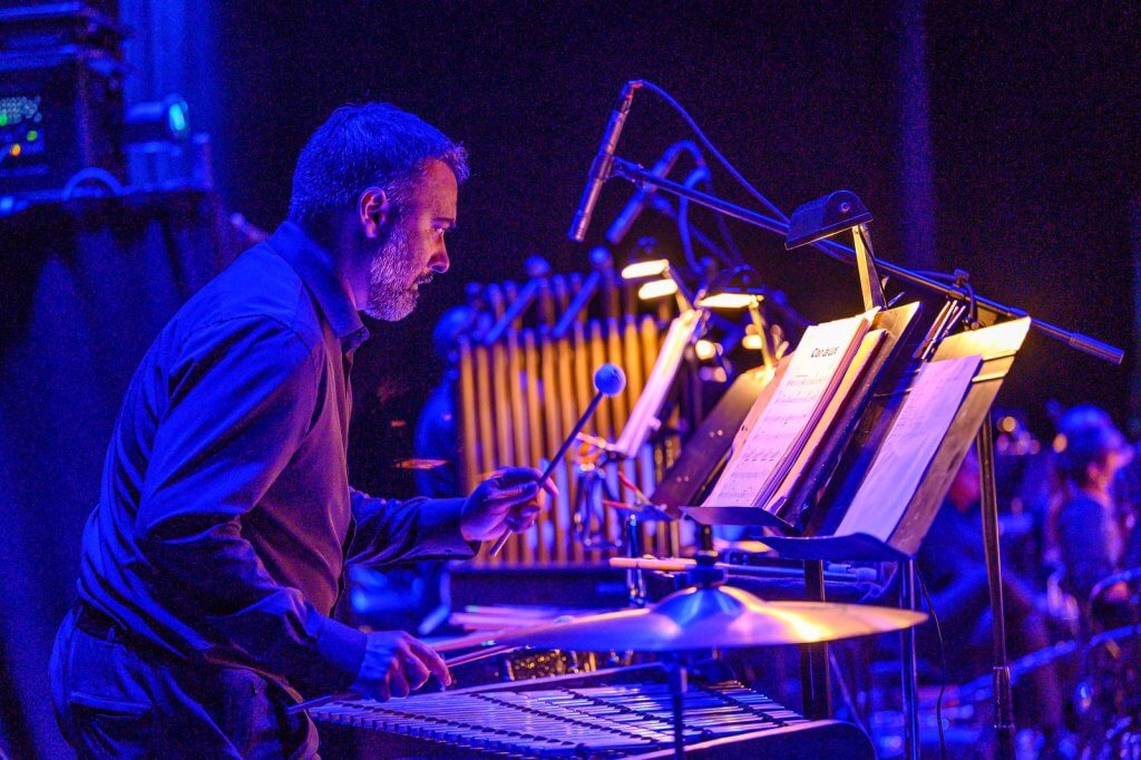 Un músico tocando el vibráfono en un escenario con poca luz, rodeado de partituras e iluminado por las luces del escenario
