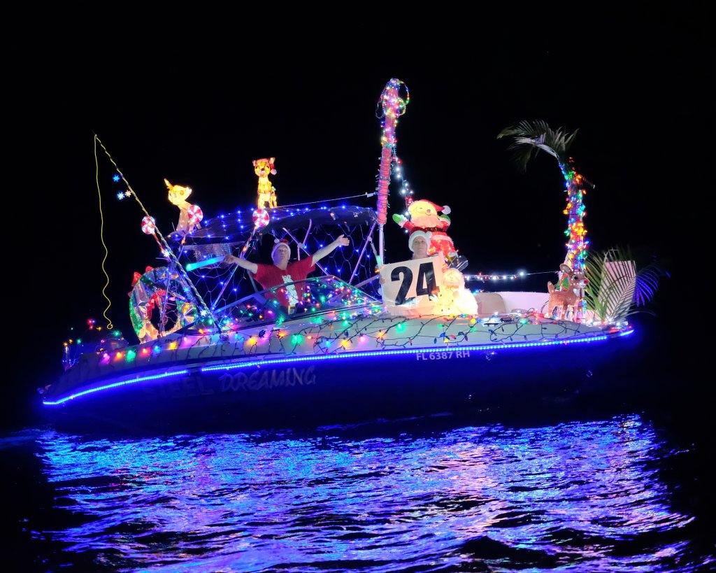 Ein hell geschmücktes Boot mit bunten Lichtern, festlichen Figuren und einem großen Weihnachtsmann nimmt an einer nächtlichen Bootsparade teil und erzeugt lebhafte Reflexionen auf dem Wasser.