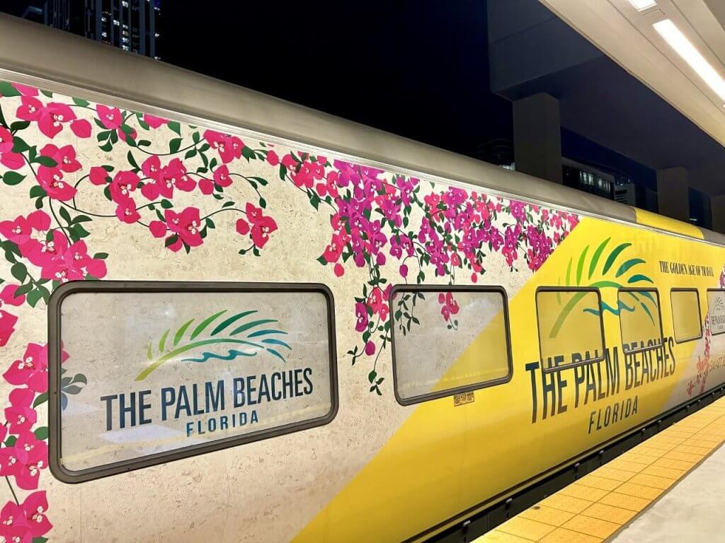 A train car adorned with vibrant pink bougainvillea flowers and palm tree graphics, promoting The Palm Beaches, Florida