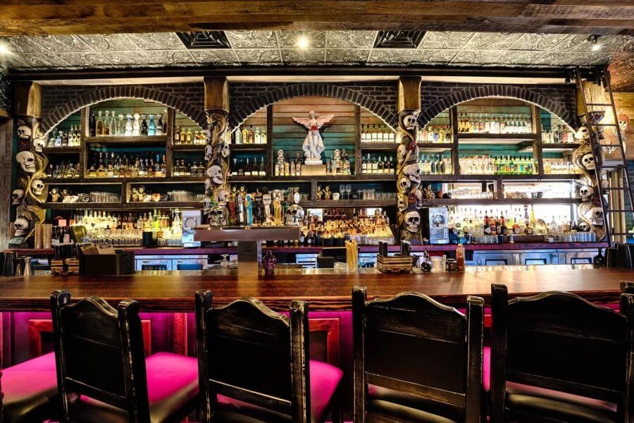 A dimly lit bar with an arched ceiling, featuring a large collection of bottles on shelves and a bar counter with pink stools