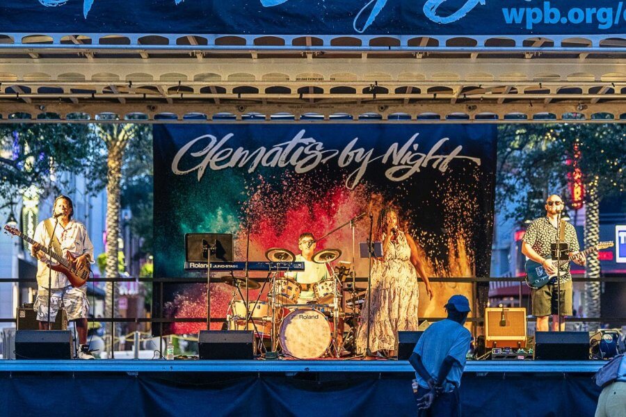 Un animado concierto al aire libre con una banda muy variada que tocará bajo una colorida pancarta que reza 'Clematis by Night'