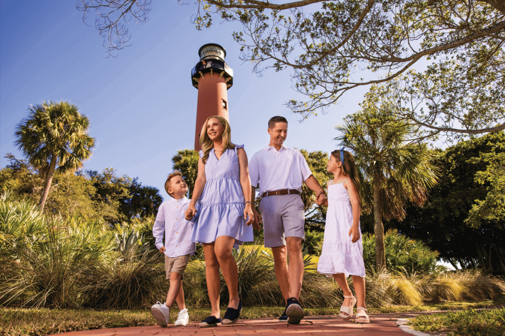 Family walking hand-in-hand near Jupiter Inlet Lighthouse surrounded by lush greenery