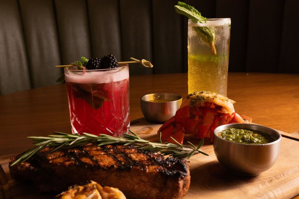 A beautifully presented steak dinner with grilled lobster tail, red cocktail, mint julep, and various sauces on a wooden board
