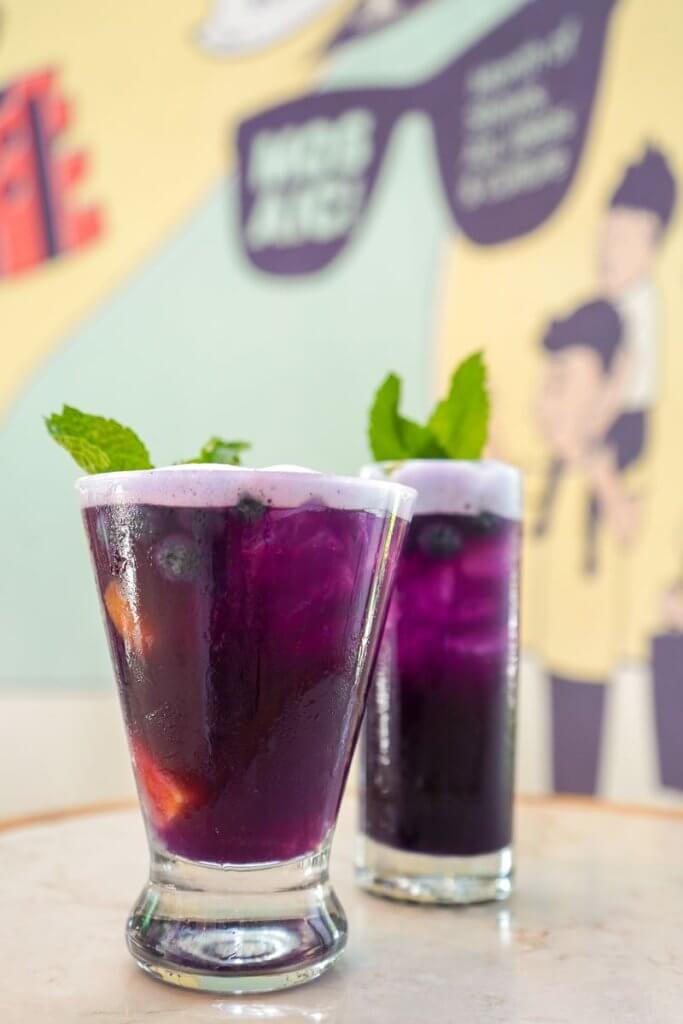 Um coquetel roxo vibrante decorado com folhas de hortelã fresca e fatias de frutas, servido em copos altos em uma mesa com um mural colorido ao fundo