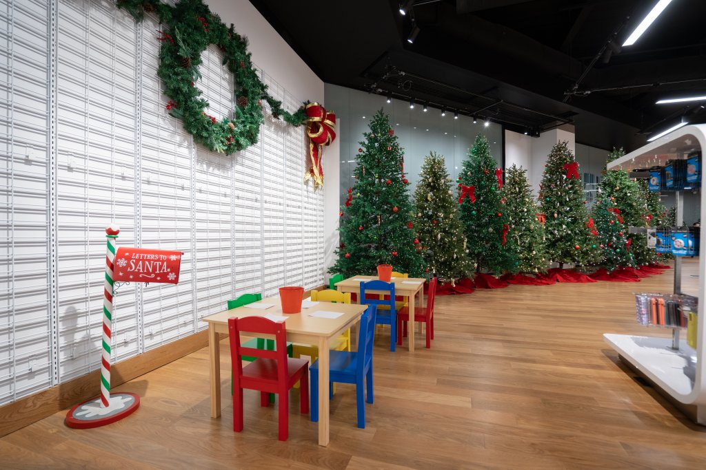 Eine festliche Einrichtung mit einer "Briefe an den Weihnachtsmann"-Station mit kleinen Tischen und bunten Stühlen für Kinder, umgeben von geschmückten Weihnachtsbäumen und Kränzen, die eine fröhliche und einladende Atmosphäre schaffen