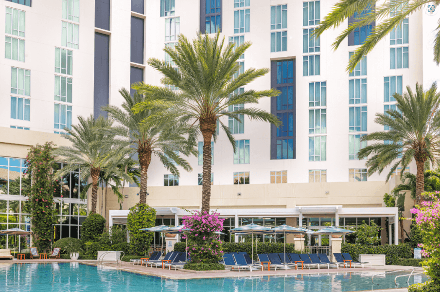 A serene resort pool area surrounded by palm trees and colorful flowers, with a modern high-rise building in the background