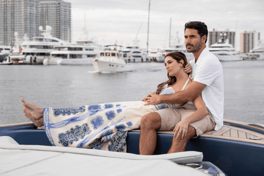 A couple enjoys a serene moment on a luxury yacht, with the woman wearing a blue and white patterned dress and the man in a white shirt and shorts, as they gaze out at the bustling marina filled with yachts and high-rise buildings in the background