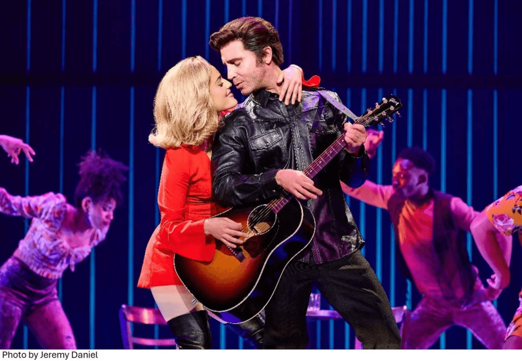 Eine leidenschaftliche musikalische Darbietung mit einem Mann in einer schwarzen Lederjacke, der eine Akustikgitarre spielt, während ihm eine Frau in einem roten Kleid ins Ohr singt, umgeben von einem lebhaften, tanzenden Refrain