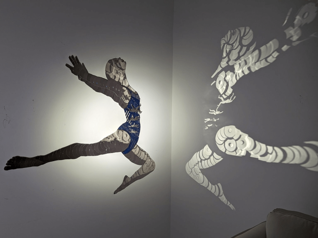 Eine auffällige Wandinstallation mit zwei miteinander verbundenen Silhouetten einer Tänzerin im Sprung, von denen eine in Blau und die andere in Weiß gehalten ist, wodurch ein fesselndes Spiel von Licht und Schatten entsteht