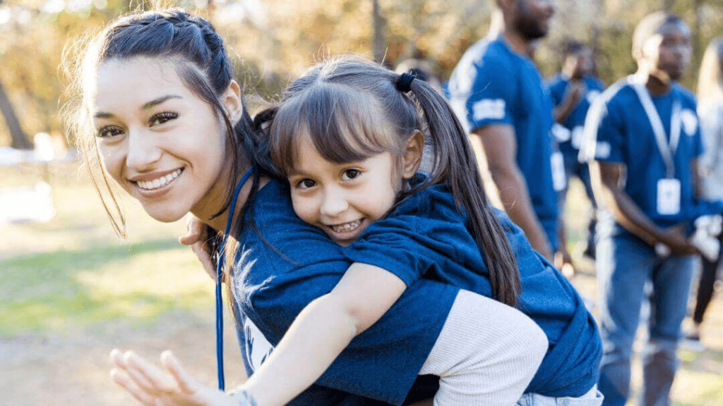 Una mujer sonriente lleva a una niña alegre a cuestas en un parque, rodeada por un grupo de personas que visten camisetas azules