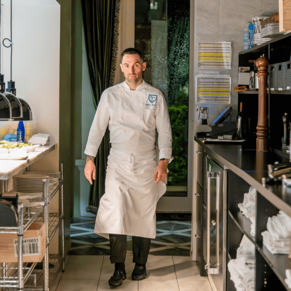 Un chef vestido con un uniforme blanco se encuentra en una cocina profesional equipada con utensilios de acero inoxidable y estantes perfectamente ordenados