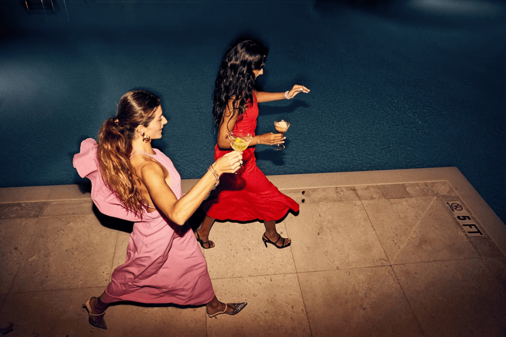 Duas mulheres em vestidos vibrantes dançam em uma piscina à noite, segurando drinques