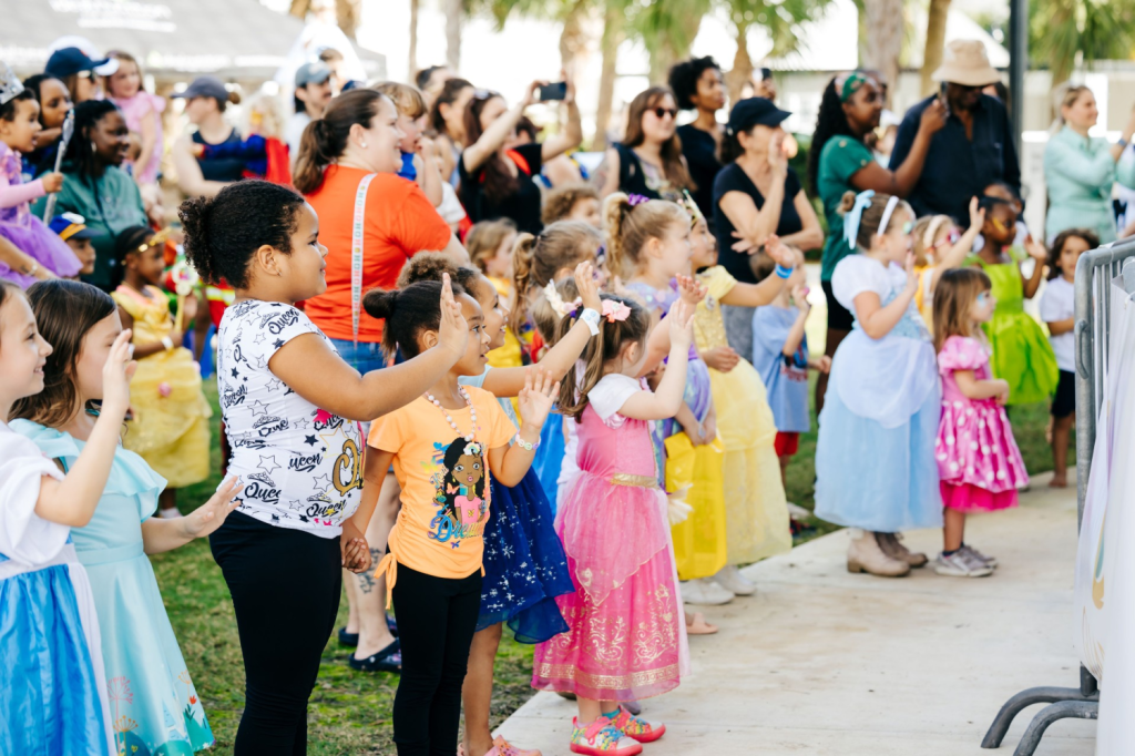 Une foule animée d'enfants vêtus de costumes de princesse colorés salue avec enthousiasme un défilé, entourés d'adultes qui les acclament.