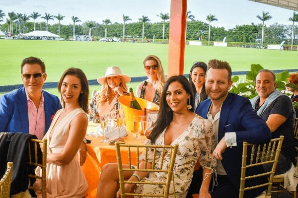 A group of stylishly dressed friends enjoys a sunny day at a polo match, sipping champagne and posing for a photo