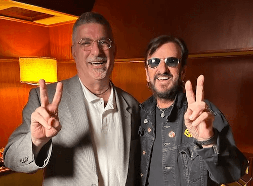 Two men, one in a suit and the other in a leather jacket, flash peace signs and smile in a warmly lit room